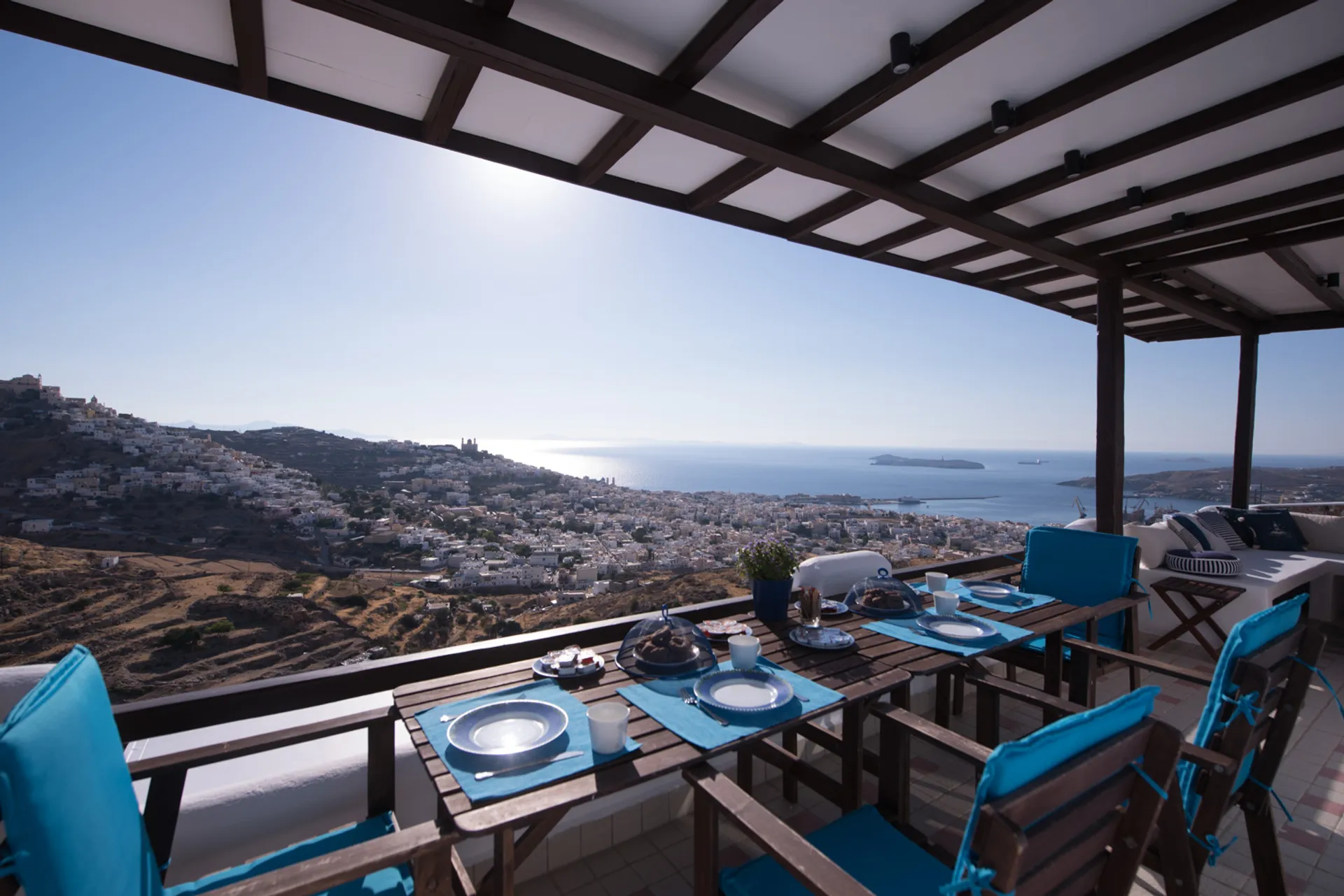 A set dining table with blue placemats on a shaded terrace overlooking the panoramic cityscape of Ermoupolis and the Aegean Sea.