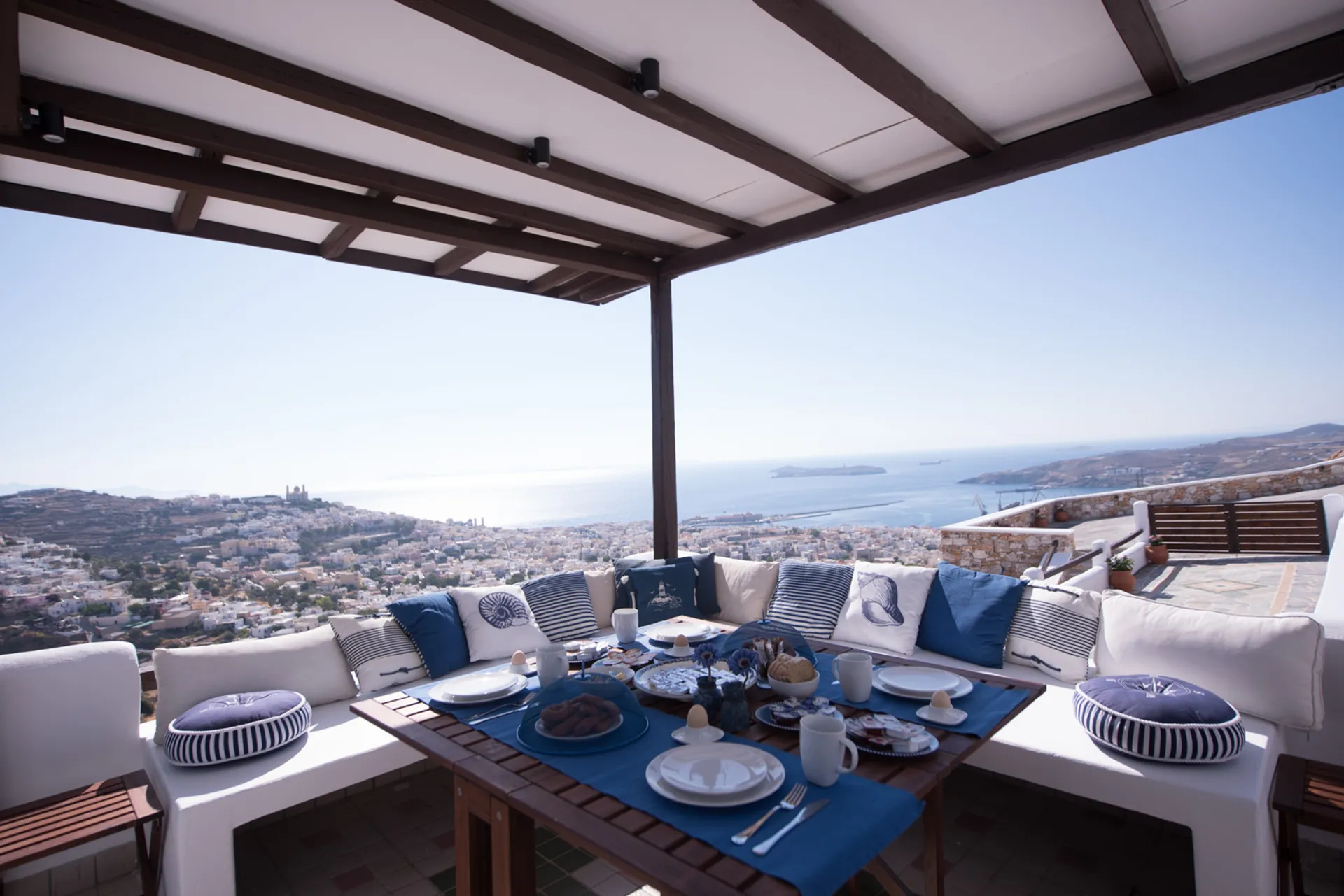 A luxury outdoor breakfast setting with nautical-themed blue and white cushions on a shaded terrace overlooking the panoramic landscape of Ermoupolis and the sea.